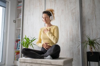 Yetişkin bir beyaz kadın milenyum kulaklıklarını internet rehberli meditasyon için kullanıyor. Evde gözleri kapalı zihin yogası yapıyor. Gerçek insanlar uzay özbakım kavramını taklit ediyorlar.