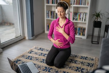 Yetişkin bir beyaz kadın milenyum kulaklığı ve dizüstü bilgisayarı kullanarak online meditasyon için bilinçlilik yogası yapıyor. Evdeki gözler kapalı gerçek insanlar mekanı kopyalıyor.