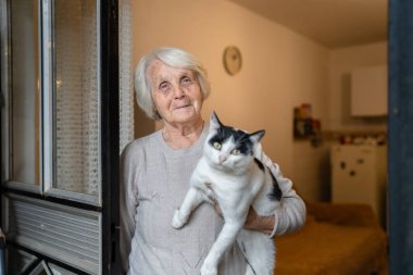 Yaşlı, yaşlı, yaşlı, beyaz, yaşlı bir kadın, evde tek başına duruyor. Belden yukarı portre, kameraya bak. Kedi evcil hayvanı, bunama hastalığı ve depresyon konsepti.