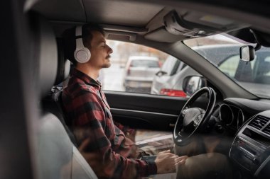 Genç, beyaz bir erkek, arabada kulaklıkla oturuyor. Rehberli meditasyon gevşemesini dinliyor. Farkındalık çalışıyor, müzik dinliyor ya da podcast yapıyor.