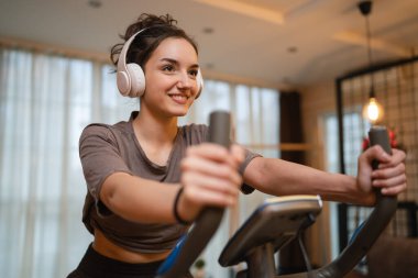 Evde kulaklıklarla bisiklet egzersizi yapan beyaz bir kadın var. Gerçek insanlar uzay sağlığı ve spor konseptini taklit ediyor. Mutlu bir gülümseme.