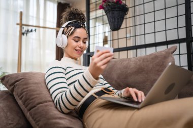 İnternetten alışveriş yapan bir kadının kredi kartı ve dizüstü bilgisayarı var. Evde genç yetişkin bir kadının evdeki kanepe yatağında otururken internetten alışveriş yaptığını görüyorlar.