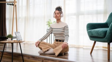 Yetişkin bir beyaz kadın milenyum kulaklıklarını internet rehberli meditasyon için kullanıyor. Evde gözleri kapalı farkındalık yogası yapıyor. Gerçek insanlar kişisel bakım kavramını taklit ediyor.