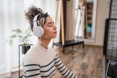 Yetişkin bir beyaz kadın milenyum kulaklıklarını internet rehberli meditasyon için kullanıyor. Evde gözleri kapalı farkındalık yogası yapıyor. Gerçek insanlar kişisel bakım kavramını taklit ediyor.