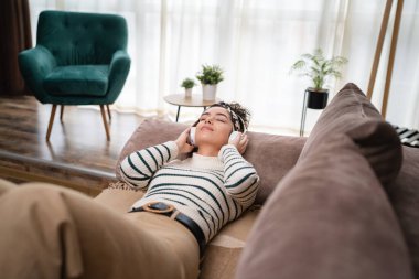 Yetişkin bir beyaz kadın milenyum kulaklıklarını internet rehberli meditasyon için kullanıyor. Düşünce yogası yapıyor. Evdeki yatakta gözleri kapalı.