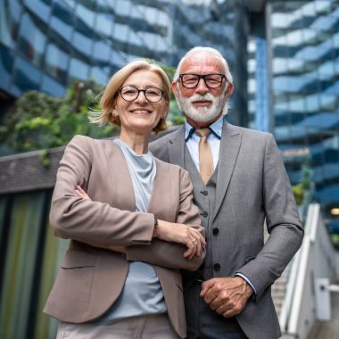 Takım elbise giymiş kıdemli bir adamın portresi ve olgun sarışın bir kadın iş ortağı olarak karı koca arkadaşları ya da iş arkadaşları modern binanın önünde duruyor.
