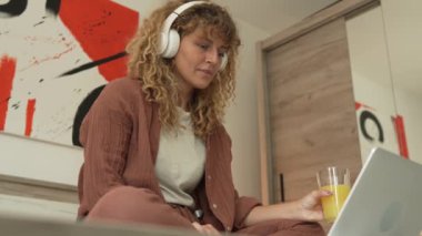 Kıvırcık saçlı beyaz bir kadın evde oturup dijital tablet ve kulaklık kullanarak film ya da dizi izliyor ya da mutlu bir gülümseme videosu çekiyor.