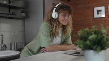 Kıvırcık saçlı beyaz bir kadın evde oturup dijital tablet ve kulaklık kullanarak film ya da dizi izliyor ya da mutlu bir gülümseme videosu çekiyor.