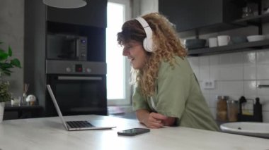 Kıvırcık saçlı beyaz bir kadın evde oturup dijital tablet ve kulaklık kullanarak film ya da dizi izliyor ya da mutlu bir gülümseme videosu çekiyor.
