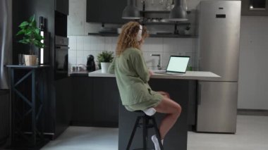 Kıvırcık saçlı beyaz bir kadın evde oturup dijital tablet ve kulaklık kullanarak film ya da dizi izliyor ya da mutlu bir gülümseme videosu çekiyor.