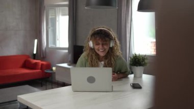 Kıvırcık saçlı beyaz bir kadın evde oturup dijital tablet ve kulaklık kullanarak film ya da dizi izliyor ya da mutlu bir gülümseme videosu çekiyor.