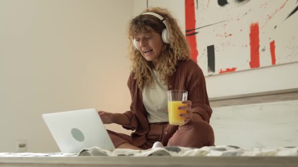 カーリーヘアを持つ1人の白人女性は 自宅で座っているデジタルタブレットとヘッドフォンを使用して 映画やシリーズをオンラインストリームで見たり ビデオコールハッピー笑顔を持っている良いタイムコピースペースを持っています — ストック動画