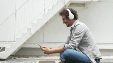 Kıvırcık saçlı beyaz bir adam modern binanın önünde oturur. Film izlemek için dijital tablet ve kulaklık kullanır. Çevrimiçi dizi podcast mutlu bir gülümseme.