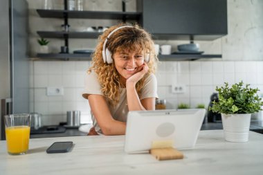 Kıvırcık saçlı beyaz bir kadın evde oturup dijital tablet ve kulaklık kullanarak film ya da dizi izliyor ya da mutlu bir gülümseme videosu çekiyor.