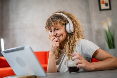 Kıvırcık saçlı beyaz bir kadın evde oturup dijital tablet ve kulaklık kullanarak film ya da dizi izliyor ya da mutlu bir gülümseme videosu çekiyor.