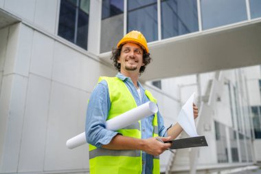 Mimar adam inşaat mühendisi ya da müfettiş kafasında koruyucu miğferle modern binanın önünde durup etrafa bakınıyor.