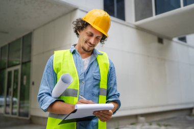 Mimar adam inşaat mühendisi ya da müfettiş kafasında koruyucu miğferle modern binanın önünde durup etrafa bakınıyor.