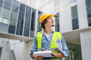 Mimar adam inşaat mühendisi ya da müfettiş kafasında koruyucu miğferle modern binanın önünde durup etrafa bakınıyor.