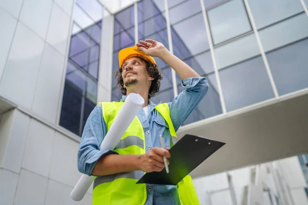 architect man construction engineer worker or inspector stand outdoors in front of modern building with protective helmet on head and documents in hands looking around