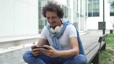Beyaz bir adam dışarıda oturur, mutlu bir şekilde gülümser, video oyunları oynar, eğlenir, cep telefonu kullanır, akıllı telefon kullanır, internetteki yetişkin erkeklerin yavaş çekimde eğlenmesini sağlarım.
