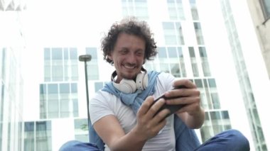 Beyaz bir adam dışarıda oturur, mutlu bir şekilde gülümser, video oyunları oynar, eğlenir, cep telefonu kullanır, akıllı telefon kullanır, internetteki yetişkin erkeklerin yavaş çekimde eğlenmesini sağlarım.