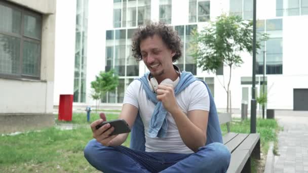 一个高加索人坐在室外刹车快乐地笑着玩电子游戏休闲活动玩着玩着手机玩着智能手机在网上玩着快乐的成年男性慢动作 — 图库视频影像
