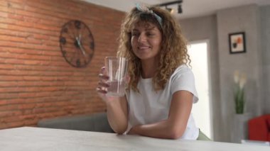 Yetişkin beyaz bir kadın evinde oturmuş bir bardak su tutarak gülümsüyor.
