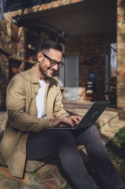 Kafkasyalı modern bir erkek balkon terasında tek başına oturuyor tişört giyiyor dizüstü bilgisayarda çalışıyor uzaktan gülüyor kendine güvenen başarı kavramını taklit ediyor.