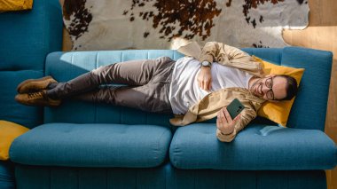 Üst görüşlü adam evde kanepede yatıyor. Cep telefonu akıllı telefonu kullan.