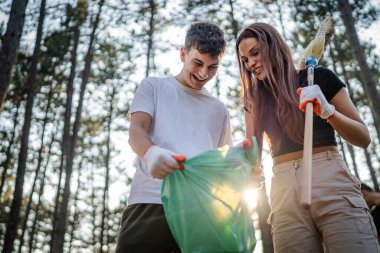 Çift ya da genç arkadaşlar Z erkek ve beyaz kadın atık plastik şişeleri ve kağıtları toplayarak güneşli bir günde çevre koruma kavramının doğasını temizlerler.