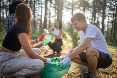 Bir grup genç arkadaş Z erkek ve beyaz kadın atık plastik şişeleri ve kağıtları toplayarak güneşli bir günde çevre koruma kavramının doğasını temizliyorlar.