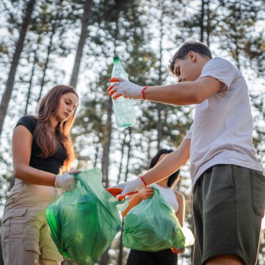 Bir grup genç arkadaş Z erkek ve beyaz kadın atık plastik şişeleri ve kağıtları toplayarak güneşli bir günde çevre koruma kavramının doğasını temizliyorlar.