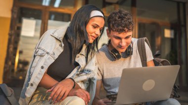 Genç çift, beyaz erkek ve dişi erkek ve kız arkadaş öğrenci erkek ve kadın okulda dizüstü bilgisayarda video izlerken güneşli bir günde açık havada mutlu bir şekilde gülümsüyor ve internette eğleniyorlar.