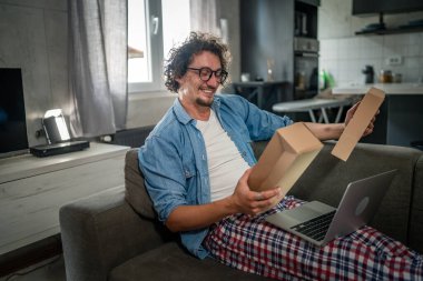 Kafkasyalı yetişkin bir erkek kanepenin üzerinde oturup sipariş ettiği postayla paketleri alır. Ya da özel hediyeli mutlu bir gülümseme. Gerçek insanlar servis kavramı.