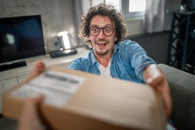 Kafkasyalı yetişkin bir erkek kanepenin üzerinde oturup sipariş ettiği postayla paketleri alır. Ya da özel hediyeli mutlu bir gülümseme. Gerçek insanlar servis kavramı.