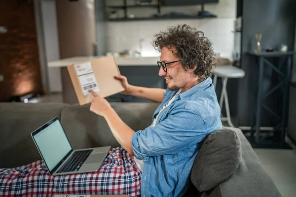 Kafkasyalı yetişkin bir erkek kanepenin üzerinde oturup sipariş ettiği postayla paketleri alır. Ya da özel hediyeli mutlu bir gülümseme. Gerçek insanlar servis kavramı.