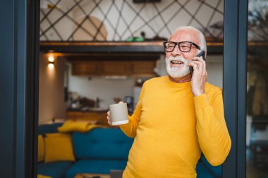 Gri sakallı, üst düzey beyaz bir erkek güler yüzlü, mutlu bir şekilde balkonda duruyor. Cep telefonunu kullanarak konuşma yeri fotokopisi çektiriyor. Günlük kahve fincanı tutuyor.