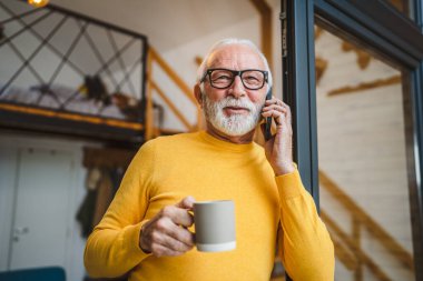 Gri sakallı, üst düzey beyaz bir erkek gün içinde balkonda duruyor. Cep telefonuyla fotokopi makinesini arıyor. Bir fincan kahveyi endişeli bir şekilde tutuyor.