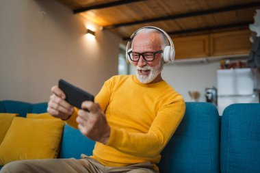 Yaşlı bir beyaz adam evinde mutlu bir şekilde oturur video oyunları oynar, eğlenir cep telefonu bulundurur, akıllı telefon alır ve eğlenceli fotokopi çekerler.