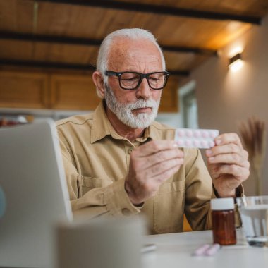 Son sınıf beyaz erkek büyükbaba evinde oturup ağrı kesici veya vitamin takviyesi paketini tutuyor. Bilgisayardan sipariş edilen dizüstü bilgisayarın yanında fotokopi odası almaya hazır.