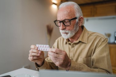 Son sınıf beyaz erkek büyükbaba evinde oturup ağrı kesici veya vitamin takviyesi paketini tutuyor. Bilgisayardan sipariş edilen dizüstü bilgisayarın yanında fotokopi odası almaya hazır.