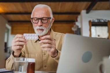 Son sınıf beyaz erkek büyükbaba evinde oturup ağrı kesici veya vitamin takviyesi paketini tutuyor. Bilgisayardan sipariş edilen dizüstü bilgisayarın yanında fotokopi odası almaya hazır.