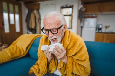 Bir beyaz erkek, son sınıf öğrencisi, evdeki kanepede oturup kağıt mendil tutarak hapşırıyor.