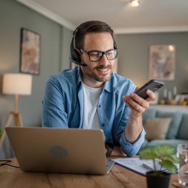 Kafkasyalı bir erkek, kafasında kulaklıkla evden çalışıyor cep telefonu ve dizüstü bilgisayar müşteri destek memuru kullanıyor.