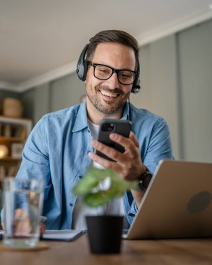 Kafkasyalı bir erkek, kafasında kulaklıkla evden çalışıyor cep telefonu ve dizüstü bilgisayar müşteri destek memuru kullanıyor.