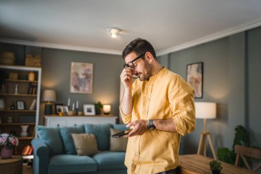 Sakallı ve gözlüklü yetişkin bir adam mutlu bir şekilde gülümseyerek ayakta duruyor cep telefonu akıllı telefonuyla mesaj atıyor ya da internette fotokopi çekiyor.