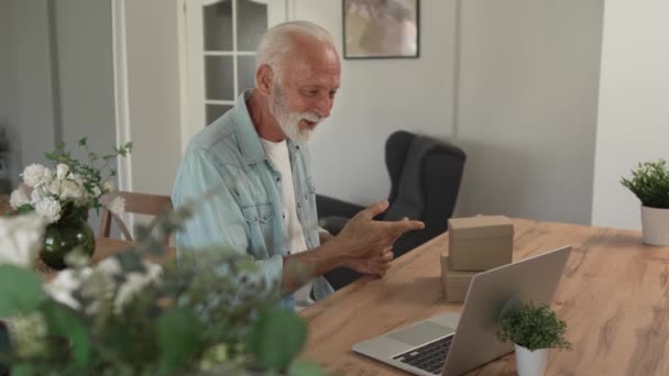 灰色の髪とひげを持つ1人のシニア男性祖父年金は オンラインビデオ通話コピースペースを持つ自宅でラップトップコンピュータの前で開いた読書カード幸せな笑顔のプレゼントを受け取ります — ストック動画