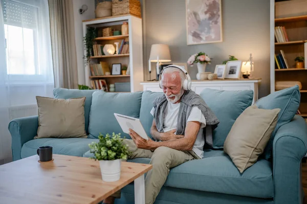 stock image One senior man old caucasian male pensioner sit at home on the sofa bed use digital tablet to watch movie or have a video call online with headphones on his head gray hair and beard