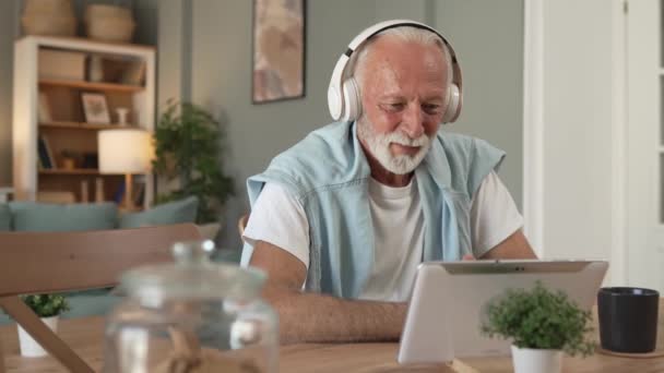 ソファベッドの自宅に座っている1歳の高齢男性年金受給者は 映画を見るためにデジタルタブレットを使用するか ヘッドグレーの髪とひげにヘッドフォンでオンラインでビデオ通話を持っています — ストック動画