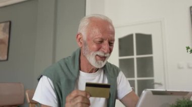 Üst düzey beyaz erkek büyükbaba evde oturuyor. İnternetten kredi kartı ve dijital tablet alışverişi yapıyor. İnternetten son sınıf öğrencisi satın almak için internet kullanıyor. 